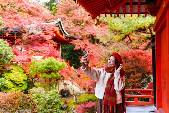 Solo female tourist taking selfie of beautiful scenic view and vibrant autumn foliage at Japanese temple in Kyoto
