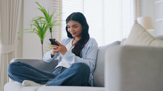Happy young asian woman relax on comfortable couch at home texting messaging on smartphone, smiling girl use cellphone, chatting online message, shopping online from home