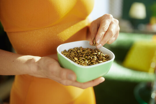 A woman in an orange top enjoys a snack of pumpkin seeds from a green bowl in a sunlit room, focusing on wellness, organic nutrition, and mindful living.