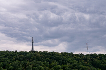 Obraz premium Petrin Lookout Tower rising above a green wooded hill under dramatic storm clouds in Prague, Czechia, with a telecommunications tower visible nearby. 