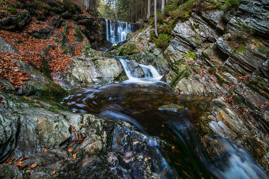 Small waterfall cascading over mossy rocks in a peaceful autumn forest with fallen orange leaves