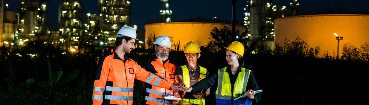 Engineers in safety gear review plans at a refinery at night, with illuminated storage tanks and industrial infrastructure in the background.