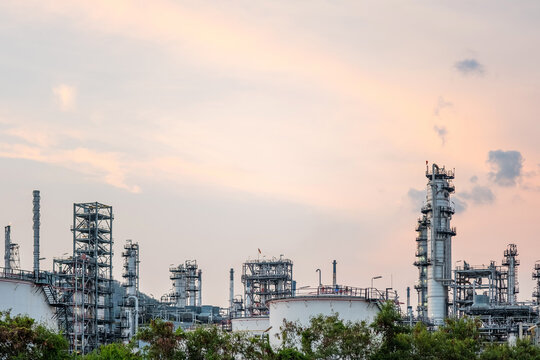 Large oil refinery with distillation towers, tanks, and piping rising above greenery under a soft evening sky