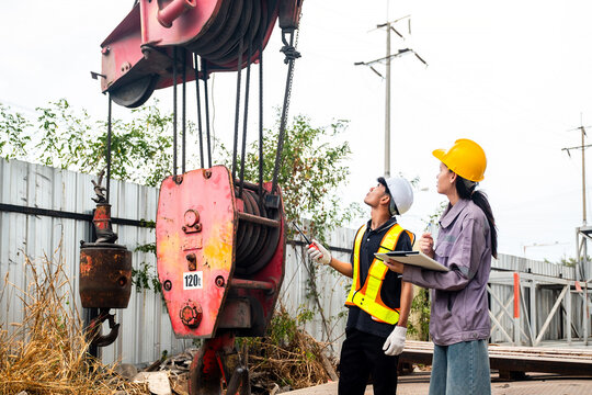Two construction workers inspect a crane hook block at a jobsite, wearing hard hats and safety gear near fencing and power lines