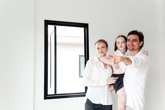 Smiling family in a bright room, the father pointing ahead while holding their child beside a window, suggesting a new home or renovation.