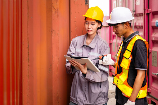 Two logistics workers in hard hats and safety vests inspect a shipping container in a cargo yard, reviewing information on a tablet during a freight operation.