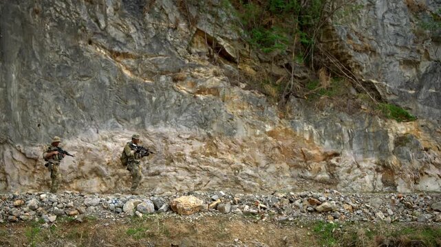 Soldiers in camouflage patrol along a rocky mountain trail beside a cliff, carrying rifles and gear in a rugged wilderness setting.