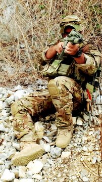 Camouflaged soldier seated on rocky terrain aims a rifle during a tactical exercise in a dry, brushy area.