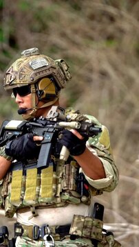 Soldier in camouflage tactical gear holds a rifle and scans the surroundings, wearing helmet, headset, body armor, and gloves in an outdoor setting