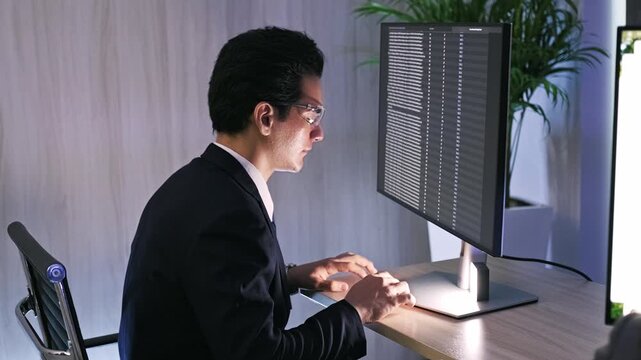 A professional works late at a desktop, focused on lines of code scrolling across a large monitor in a modern office.