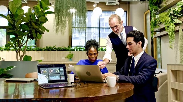 Three professionals collaborate in a plant filled modern office, reviewing analytics on laptops while discussing strategy around a wooden table.
