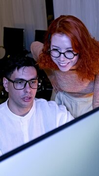 Two coworkers wearing glasses collaborate at a computer, discussing content on a large monitor in a modern office.