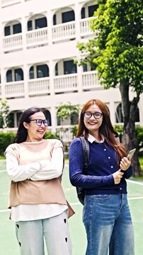 Two smiling students with backpacks laugh together on a sunny campus courtyard beside a white arched building and trees.