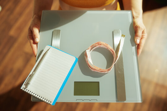 A woman in a yellow top holds a digital scale topped with a notebook and pink measuring tape, representing a commitment to weight management and fitness goals.