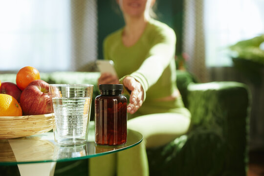 Dressed in green athletic wear, a woman reaches for water and supplements on a coffee table, highlighting a modern wellness routine that combines dietary discipline and hydration.