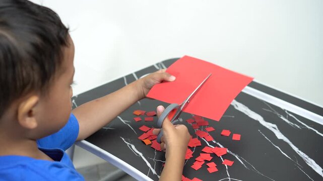 A small Asian boy is focused on learning to cut a red folded paper into small squares which will be used to stick a picture on.