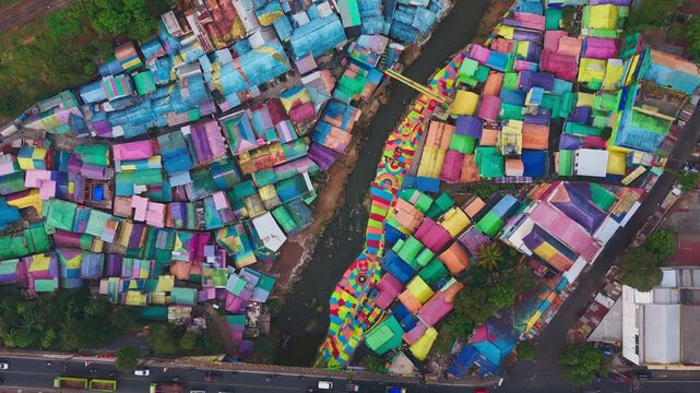Colorful rooftops dominate this vibrant slum settlement. Winding paths cut through densely packed structures. Muddy river runs alongside the crowded dwellings. Buildings show varied hues