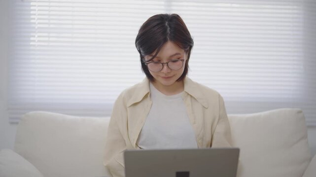 Front View of Asian Woman in Glasses Sitting on Couch and Typing on Laptop Computer in Living Room at Home