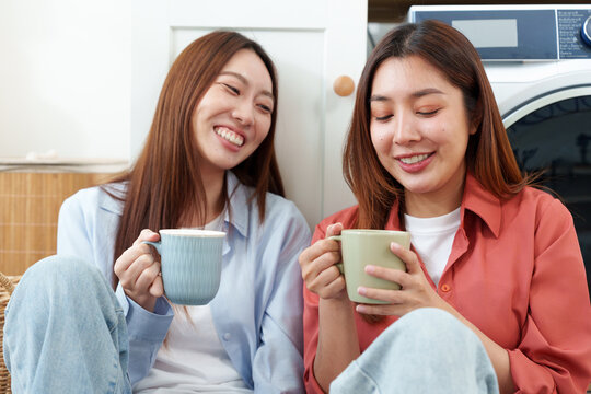 Two young happy, cheerful Asian women sitting side by side, leaning against washing machine, holding coffee mug, talking, smiling and laughing in cozy laundry room at home with joy