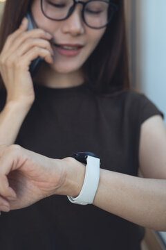 Young asian woman smiling while talking on smartphone and checking time on smartwatch, close up