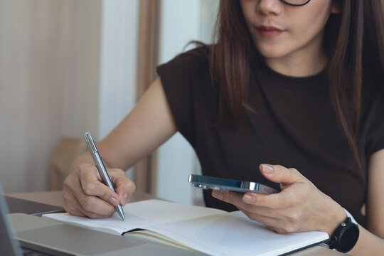 Young asian business woman using smartphone and laptop, writing note in notebook for work planning or schedule. Student studying online class , close up