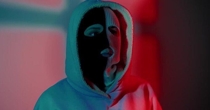 Woman in balaclava and hoodie posing under moody neon lighting. Mysterious person in face mask standing in dark studio. Concept of crime, protest or anonymity.
