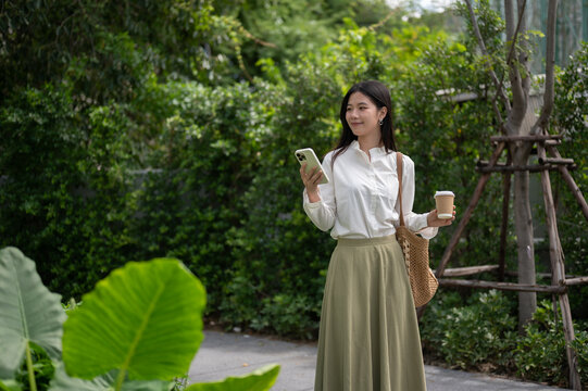 Young Asian woman smiling while holding coffee cup and smartphone while walking in green garden park