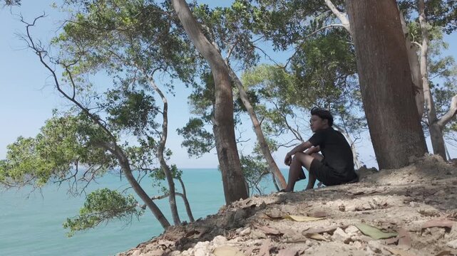 Young Asian Man Sitting with Camera Overlooking Tropical Ocean View in Kebumen, Indonesia