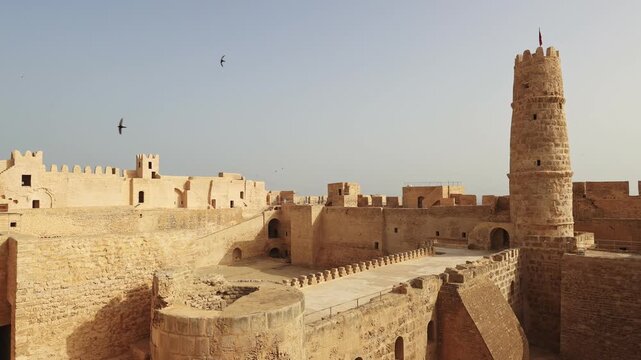 Inner courtyard of Ribat Harthama in Monastir showcasing traditional historic stone architecture and tall cylindrical watchtower bathed in golden sunlight under clear Tunisian sky. High quality 4k