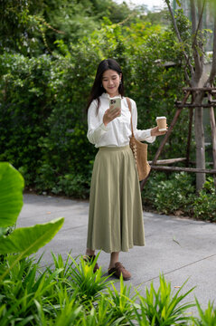 Young Asian woman smiling while using smartphone and holding takeaway coffee walking in the park.