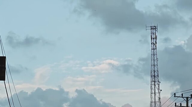 Tall communication tower standing against a cloudy sky during a late afternoon with power lines and urban infrastructure in the foreground