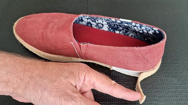 A hand points to the detached jute sole of a worn canvas shoe, highlighting damage, sole separation, and the need for repairs. Close up view of footwear wear and tear