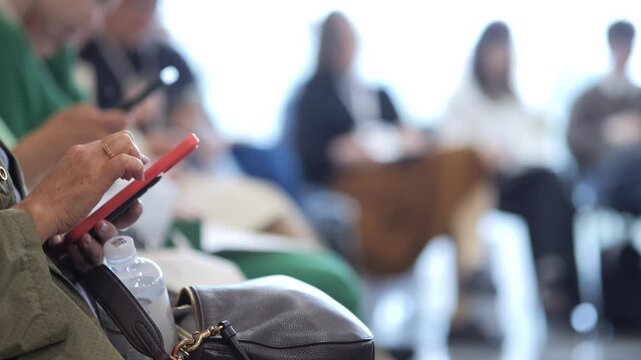 Close-up shows hand holding red tablet. Person sits in waiting area. Background features blurred attendees. Some look at devices, others appear bored or tired