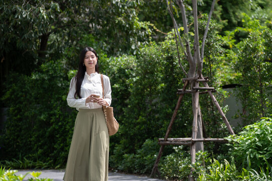 Young Asian woman holding coffee cup and enjoying a nature on a peaceful walk in green garden park.