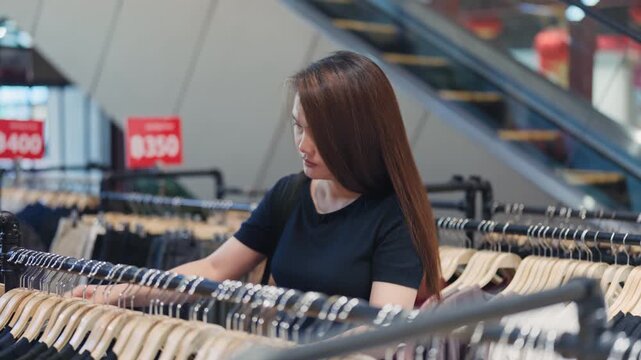 Young woman looks through garments hanging on rails with sale price signs and an escalator behind her in a store.