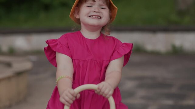 Crying child in yellow hat sitting on playground swing. Little girl wearing pink dress feeling sad outdoor. Emotion of unhappy kid playing on children equipment.