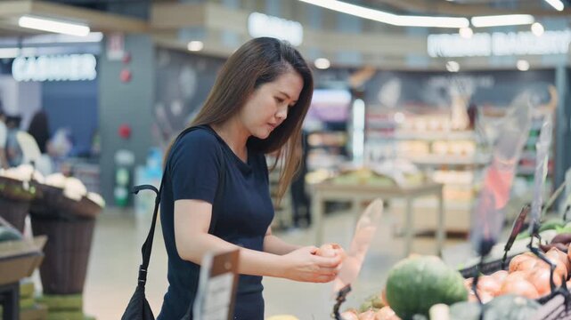 Young woman picks up a loaf of bread from a display at a supermarket bakery counter with produce nearby.