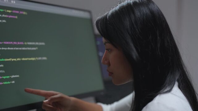 Female developer points at a line of program code on her screen while reviewing her work in a modern office.