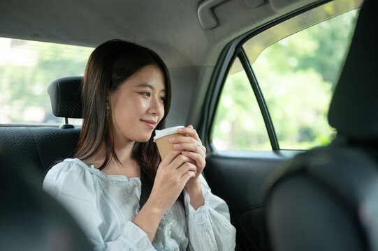 Young Asian woman holding a warm coffee enjoy drinking while relaxing in back seat of a taxi cab car
