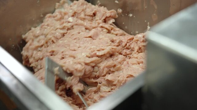 Ground meat mixture combines with poured liquid in a stainless steel mixing tank at a commercial food manufacturer.