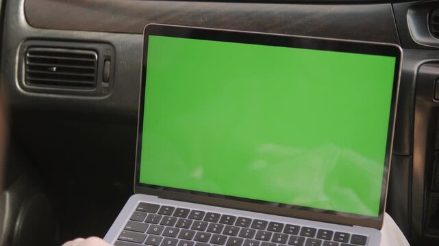Laptop with green screen sits inside car. Hands type on keyboard. View shows dashboard and air vents. Useful for travel, remote work, or digital content scenes