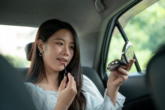 Young Asian woman applying a lipstick tint with a compact mirror in back seat during a car journey.