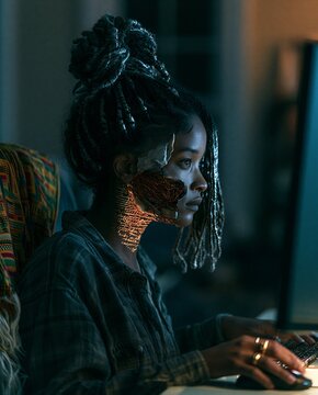 A young woman with braided hair sits in front of a computer screen, her face illuminated by its glow, suggesting deep concentration or perhaps concern