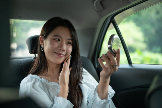 Young Asian woman applying makeup with a compact mirror while riding in back seat of a taxi cab car