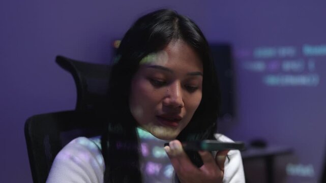 Young Asian female software developer recording a voice message on a smartphone in a dark room illuminated by computer code projections, highlighting modern IT and tech work.