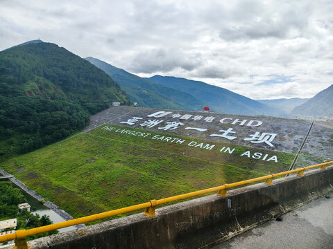 Scenic embankment dam structure with English and Chinese text "The Highest Earth Dam in Asia", green mountain range background under cloudy sky at Meijiang Dam. Yunnan-Guizhou, China. August 2, 2026.