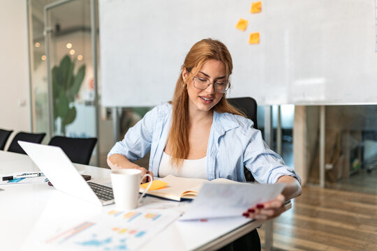 Smiling young businesswoman with vitiligo wearing glasses and blue shirt, reviewing financial documents and data reports at office desk with laptop and charts in modern workplace.