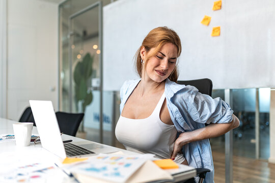 Young businesswoman with vitiligo suffering from severe lower back pain, sitting at office desk and holding her aching back while working on laptop in modern corporate workspace.