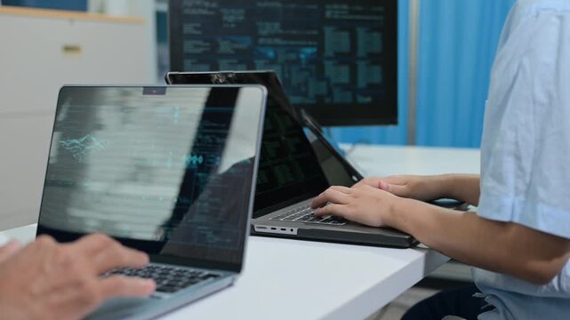Software developers collaborate at an office desk, typing code and analyzing complex data on multiple laptops and desktop displays.