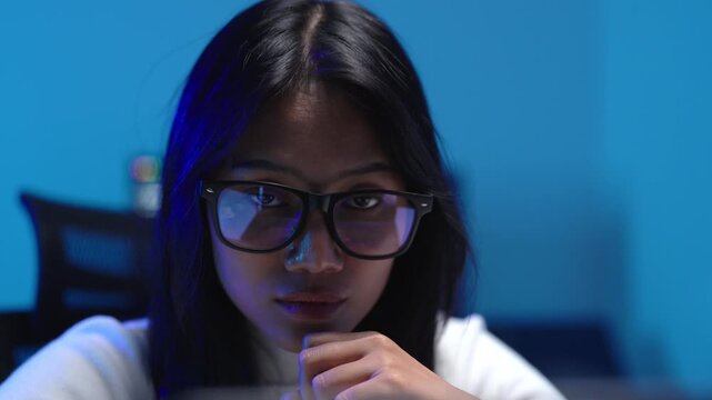 Close up of a focused Asian female programmer wearing glasses, pointing and analyzing AI generated code on a computer screen in a dark blue technology office.